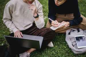 Two people sit on grass looking at laptop and one makes notes